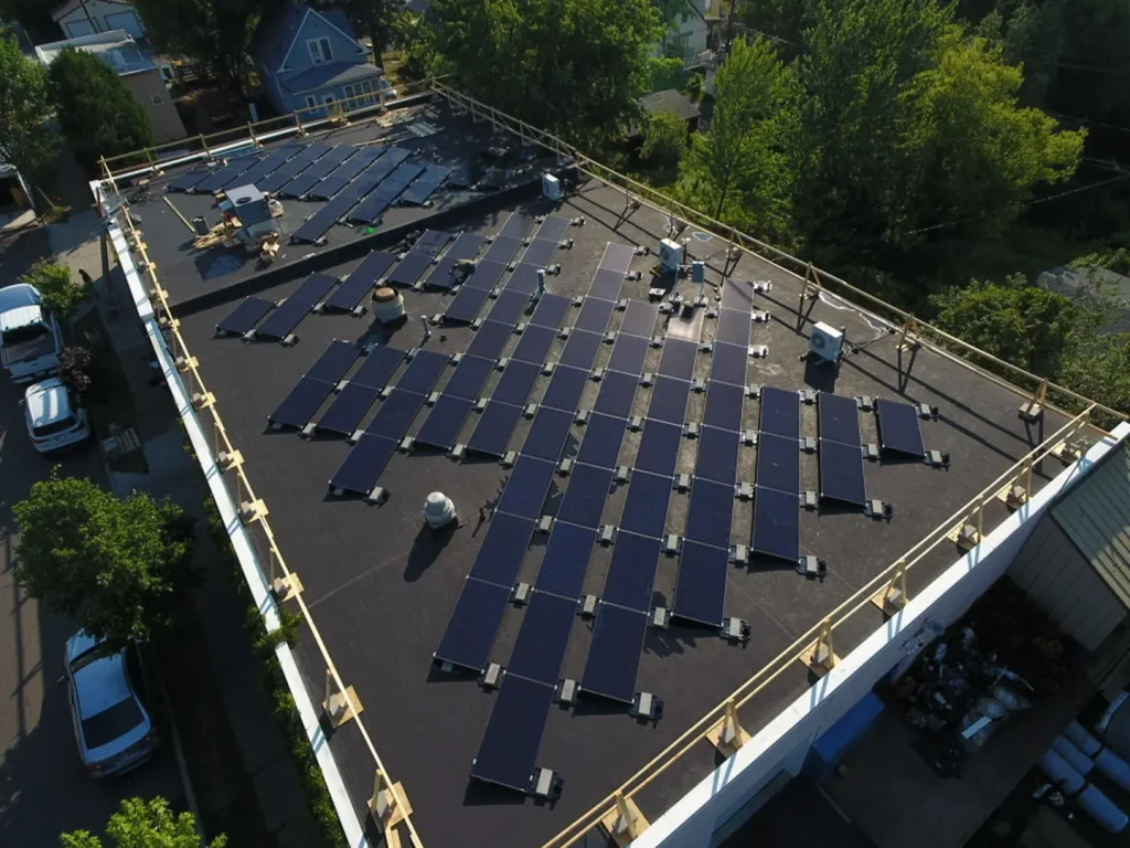 A rooftop covered with rows of solar panels, surrounded by trees and nearby houses, viewed from above in daylight.