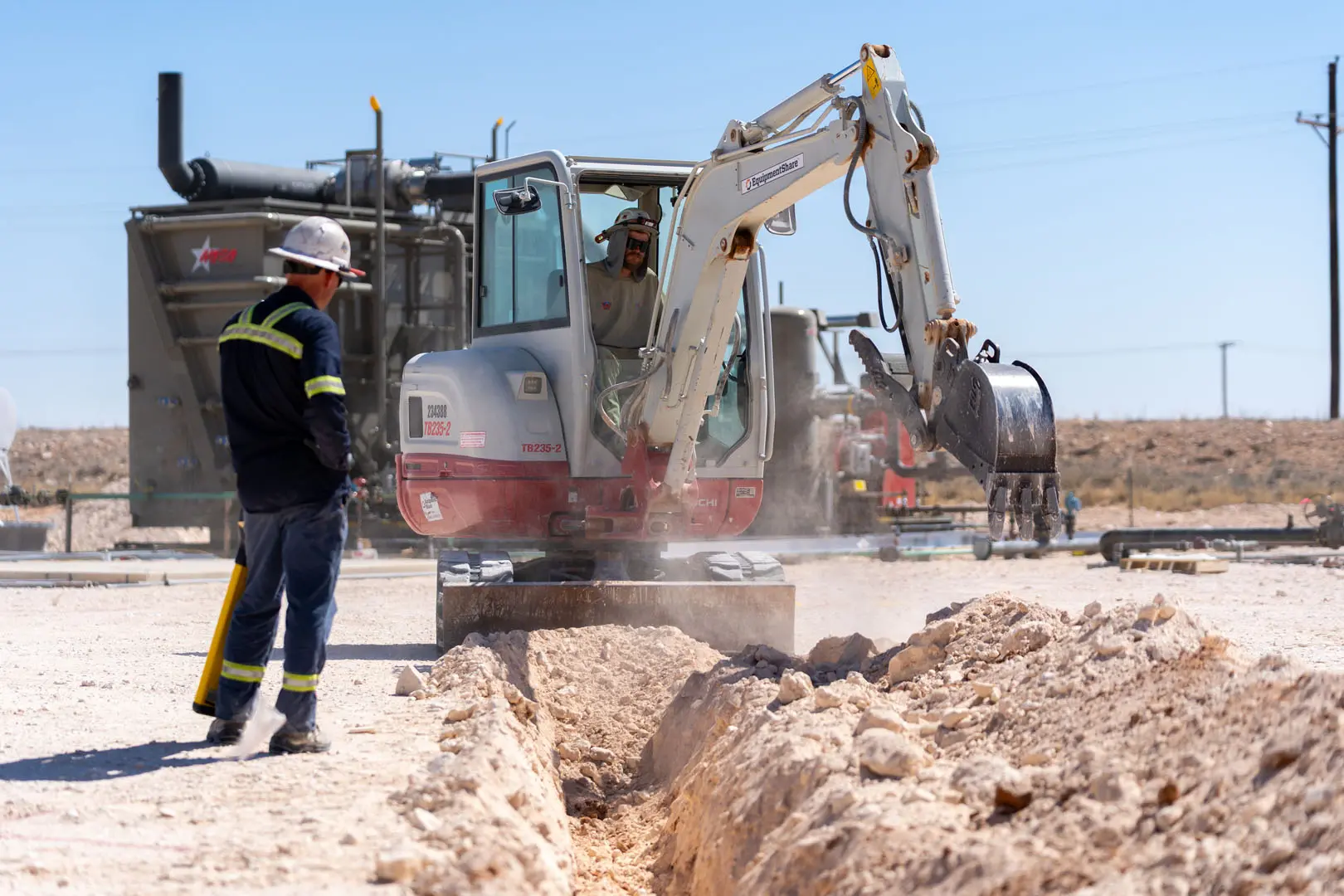 A construction worker operates a small excavator to dig a trench in rocky ground, while another worker wearing a safety vest and helmet observes nearby. Industrial equipment is visible in the background.