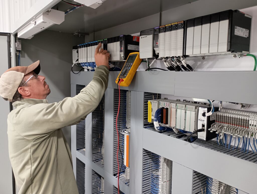 A person wearing a cap and glasses uses a tool to inspect or adjust components inside an electrical control panel, with wiring and various electronic modules visible. A multimeter hangs nearby for electrical measurements.