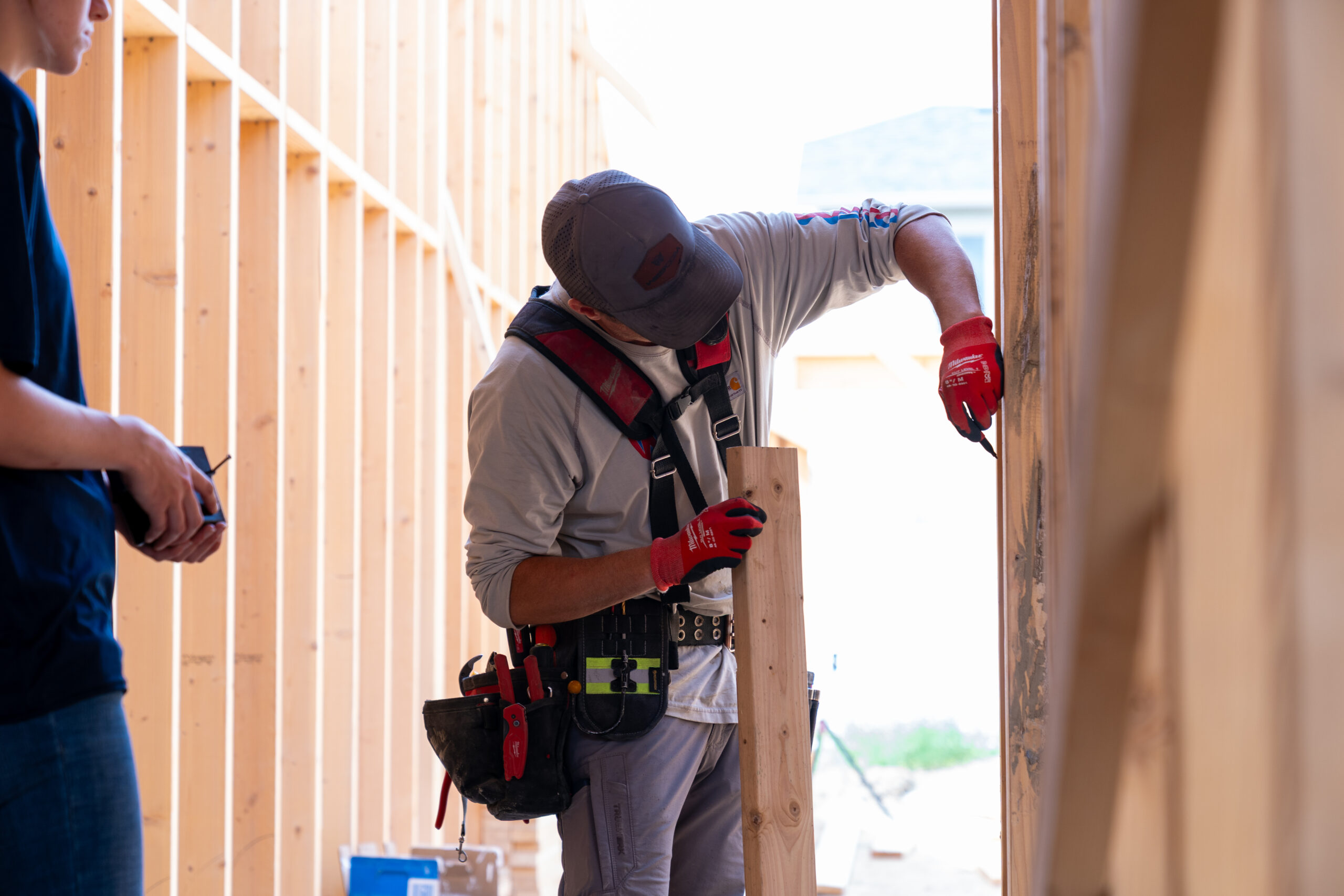 A construction worker wearing a cap, gloves, and tool belt measures a wooden beam against a wall frame at a building site, while another person stands nearby holding a phone or tool.