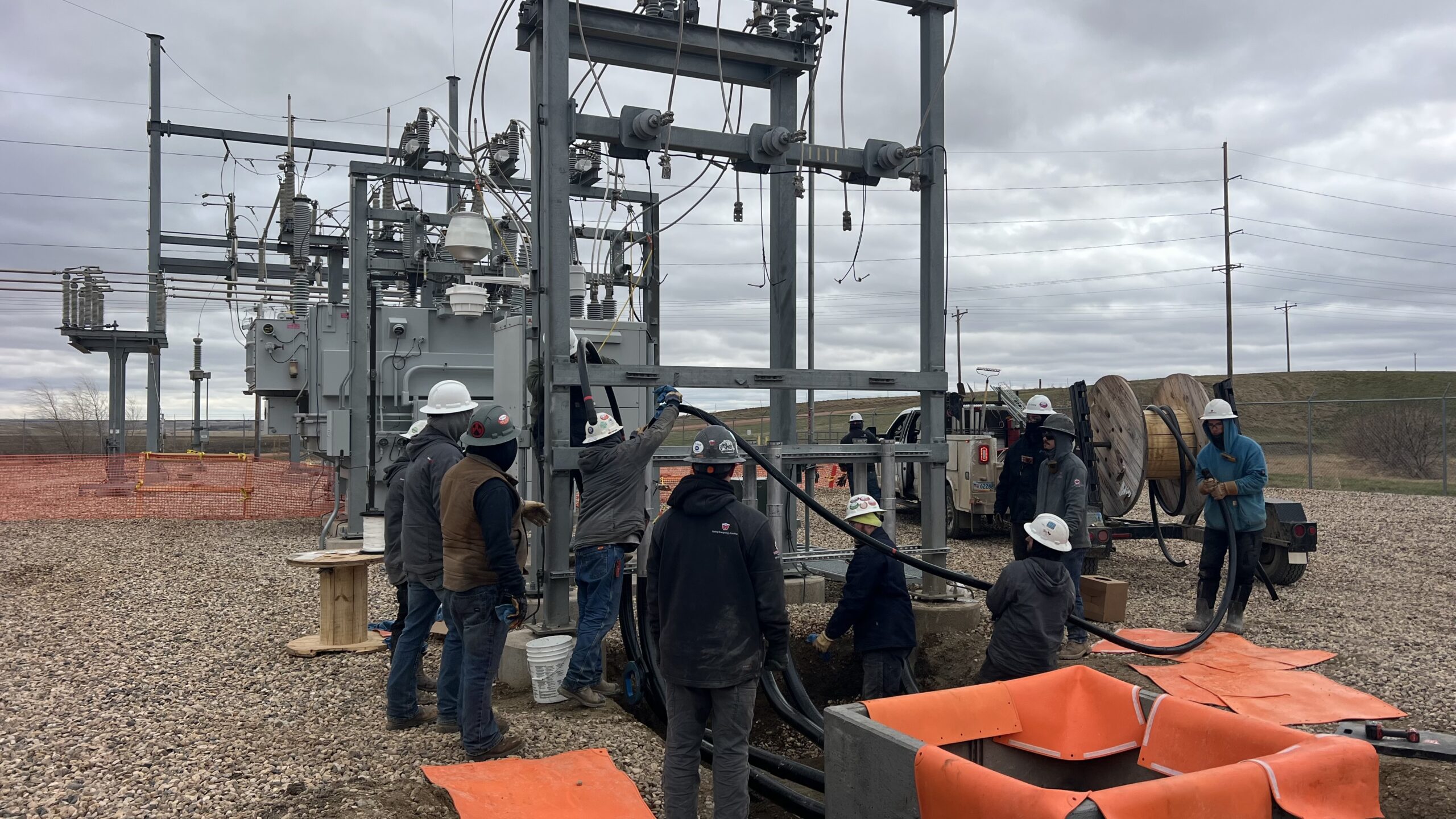 A group of utility workers in hard hats and safety gear install and inspect cables at an outdoor electrical substation on a cloudy day. Equipment, tools, and orange safety barriers are visible on the gravel ground.