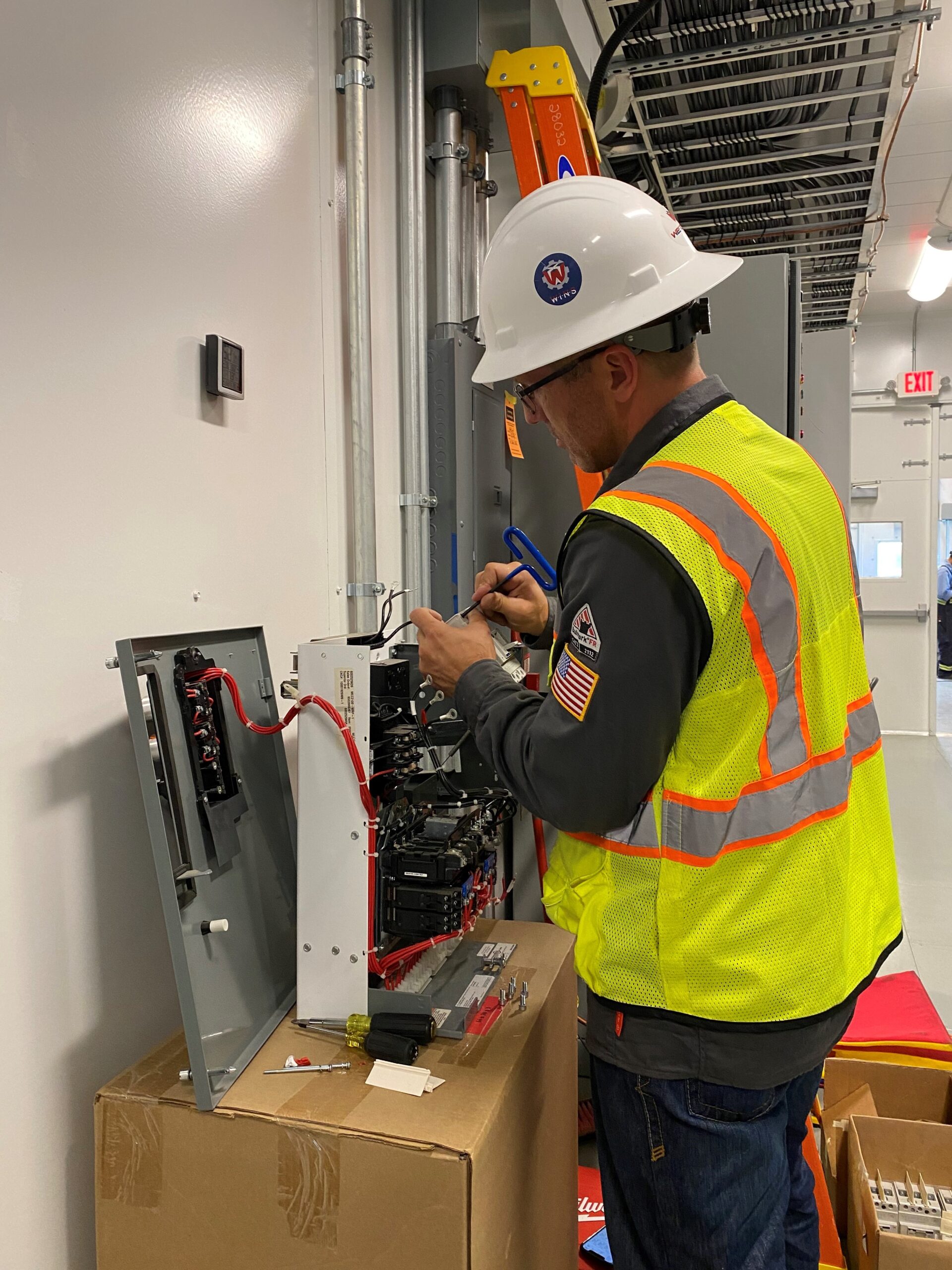 Man in safety vest and helmet doing electrical work inside control room. He is committed to safe practices. 