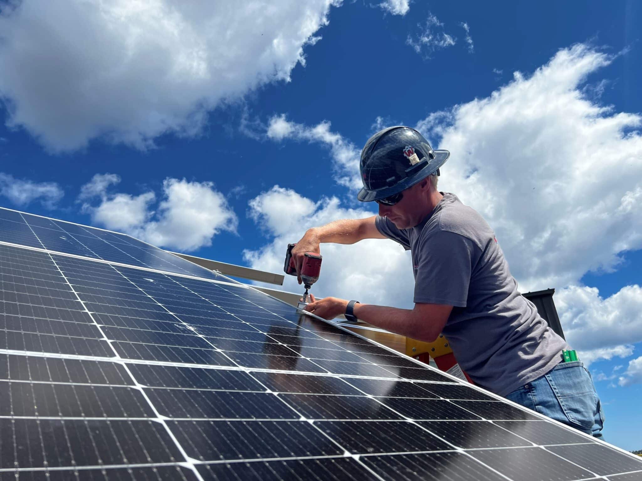 Man working on a solar array with a blue sky and clouds in the background. Solar is a great way to invest in the future.