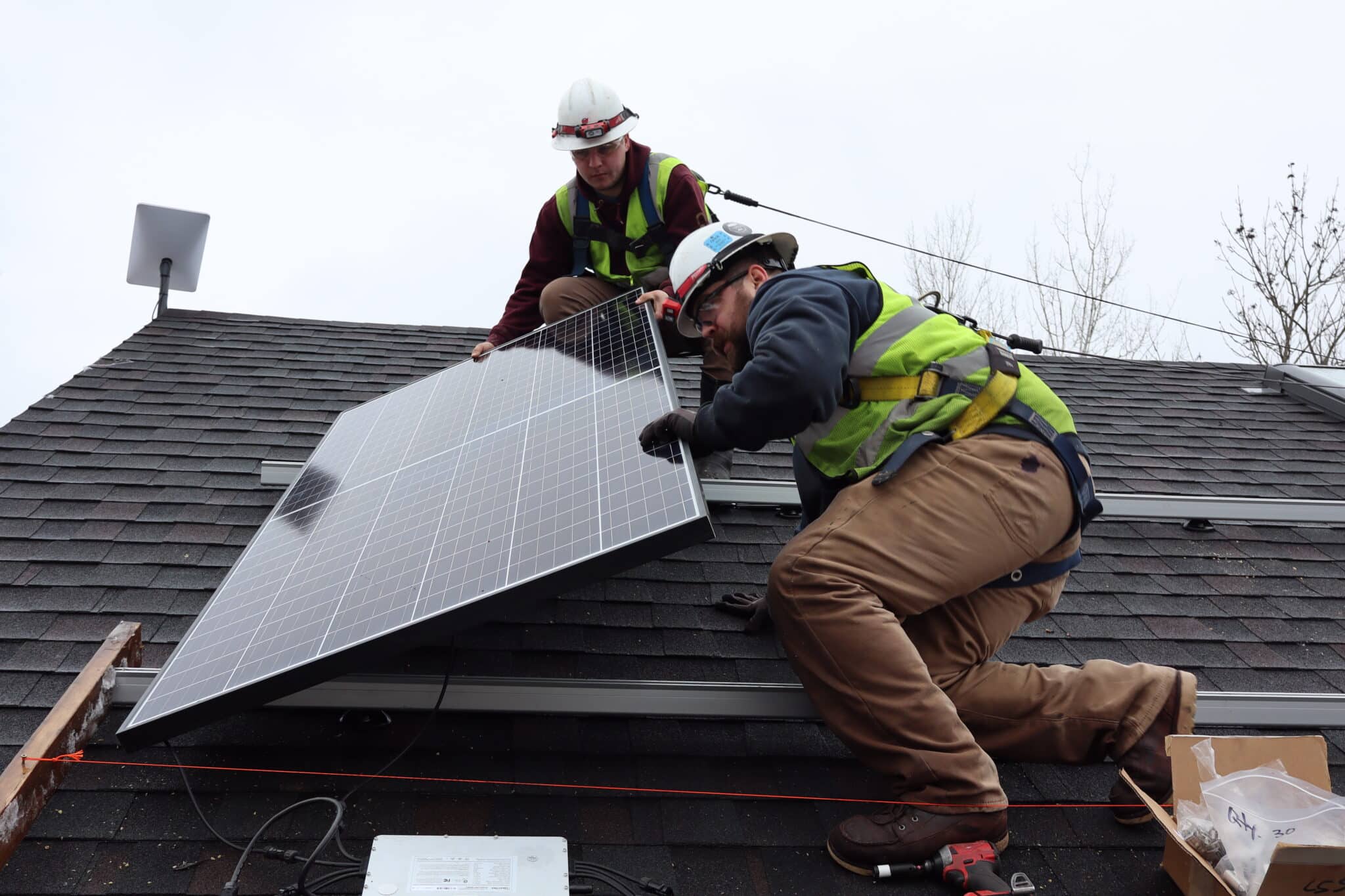 residential electricians in high vis and protective gear installing a solar panel on a gray shingle roof